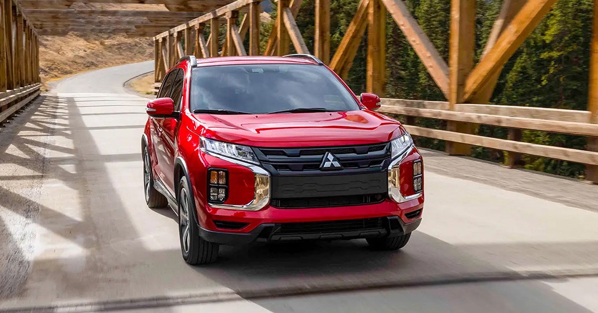 A red 2026 Mitsubishi Outlander Sport driving across a rustic bridge with a forested hillside